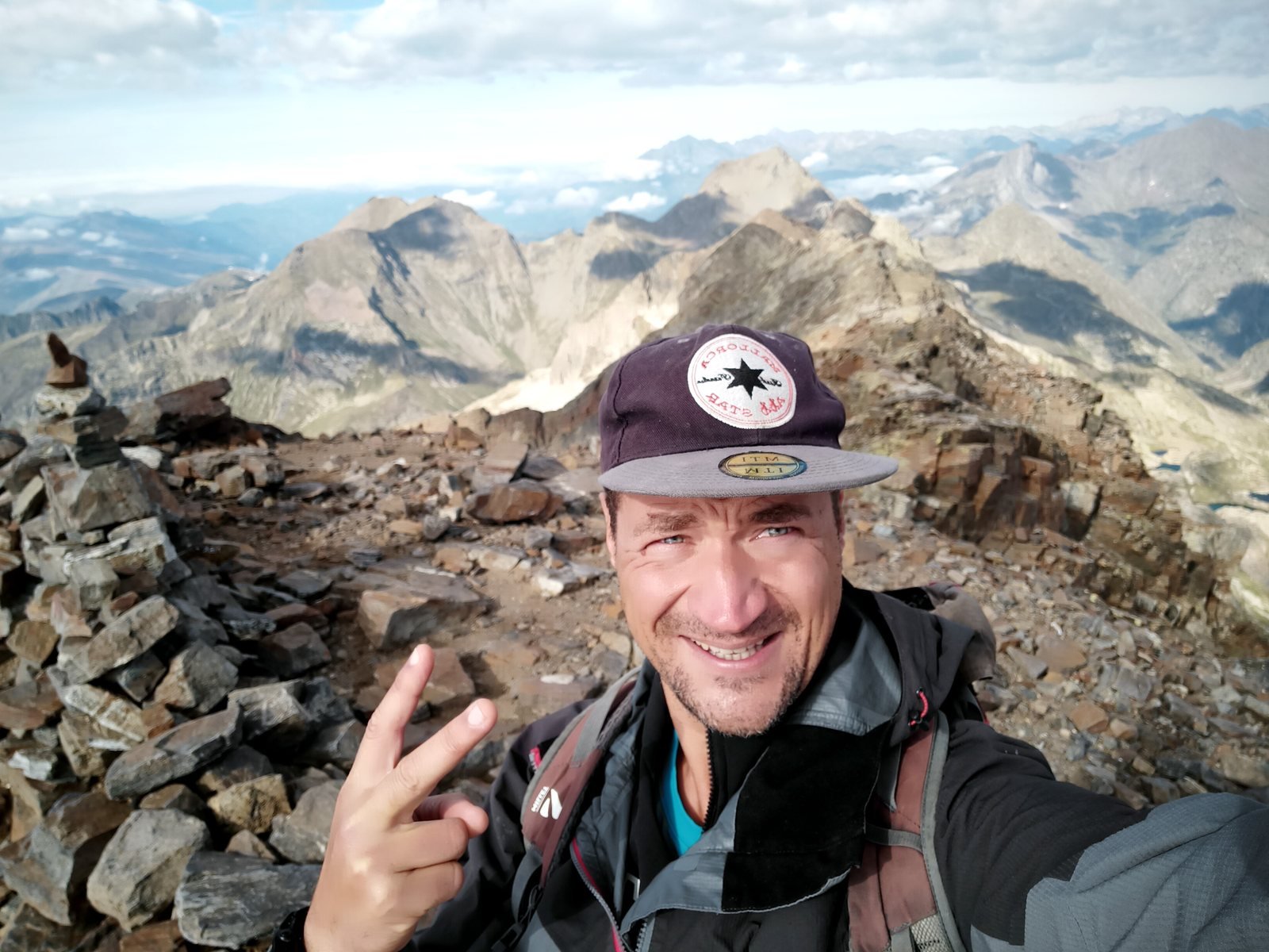 Christophe, sourire et casquette, au sommet à côté du cairn, face aux crêtes